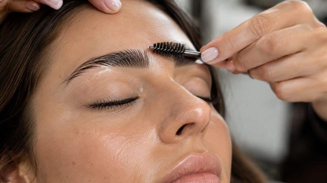 Artist brushing laminated eyebrows upward with a spoolie during a custom brow lamination